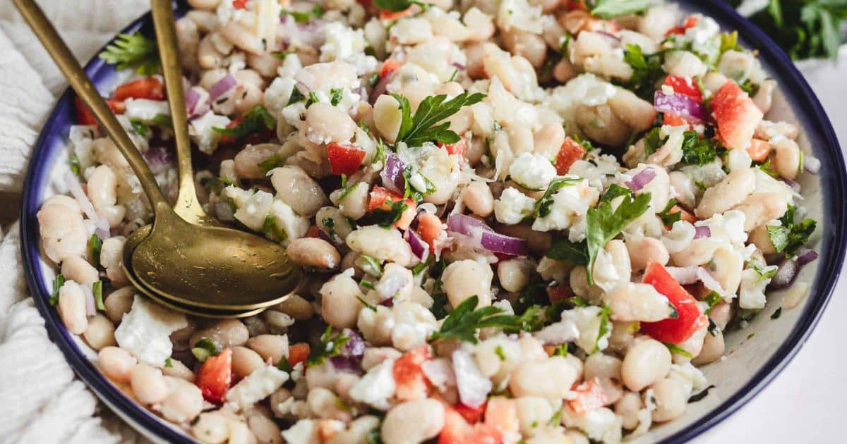 A bowl of white bean salad with red peppers, onions, parsley, and feta cheese, served with two gold spoons.