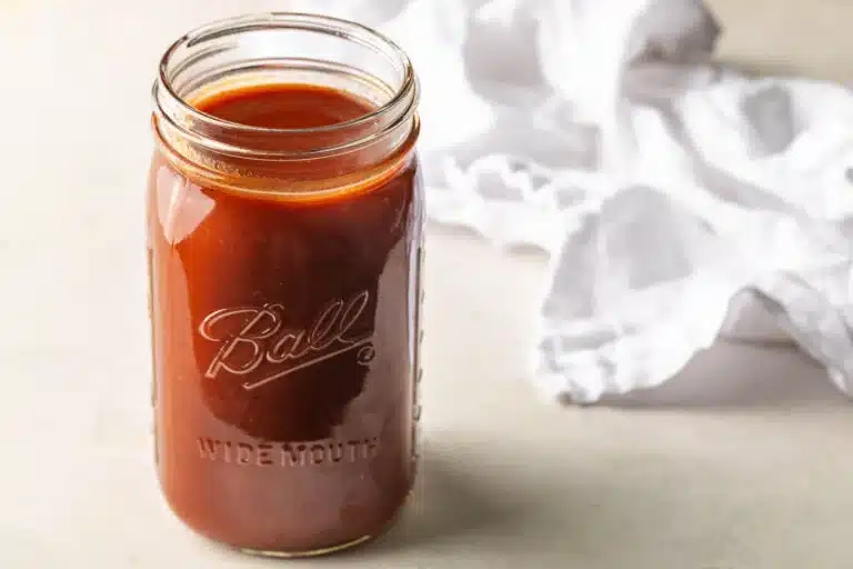 A glass jar filled with red sauce sits on a light surface next to a crumpled white cloth.