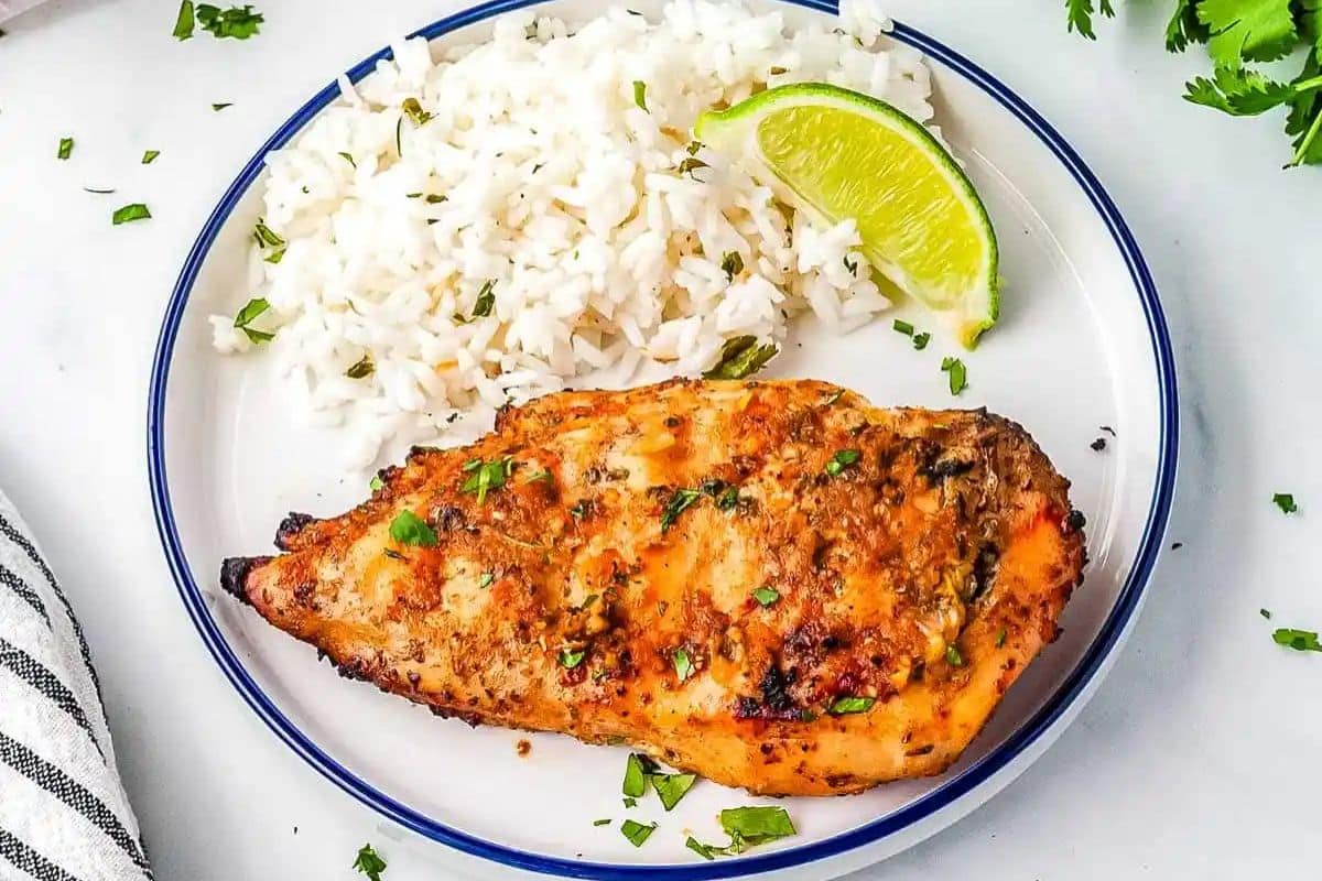 Grilled chicken breast with white rice and a lime wedge on a white plate, garnished with chopped herbs.