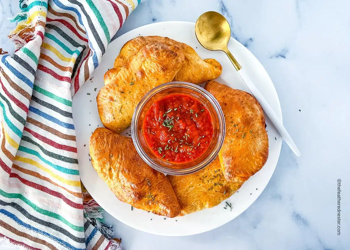 A plate of golden croissants with a bowl of red dipping sauce, next to a striped cloth and gold spoon.