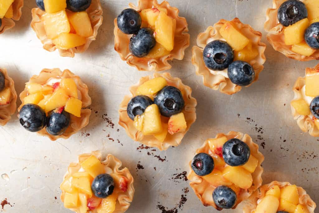 Mini tart shells filled with diced peaches and blueberries arranged on a metal baking sheet.