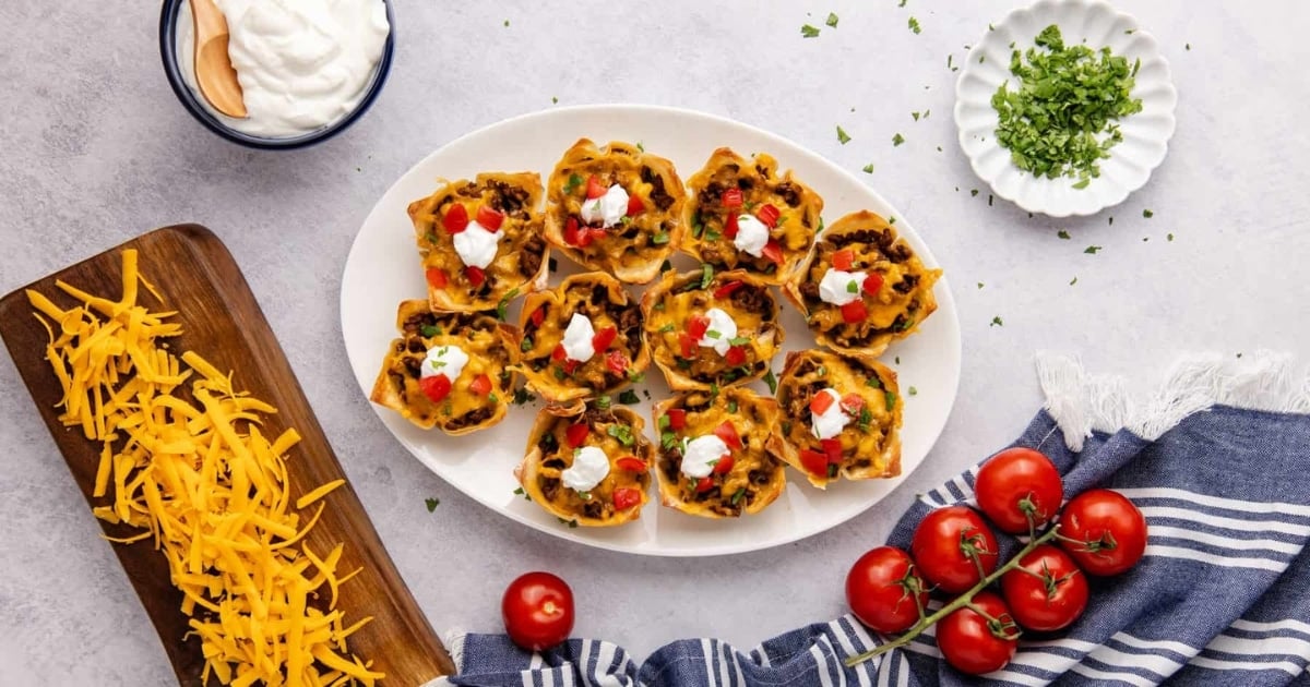 Mini taco cups topped with sour cream on a plate, surrounded by cheese, tomatoes, and fresh herbs.