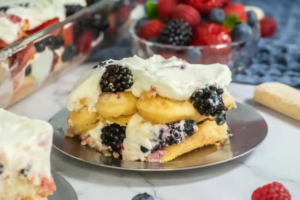 A slice of berry tiramisu with cream, blackberries, and ladyfingers on a metal plate, with berries in the background.