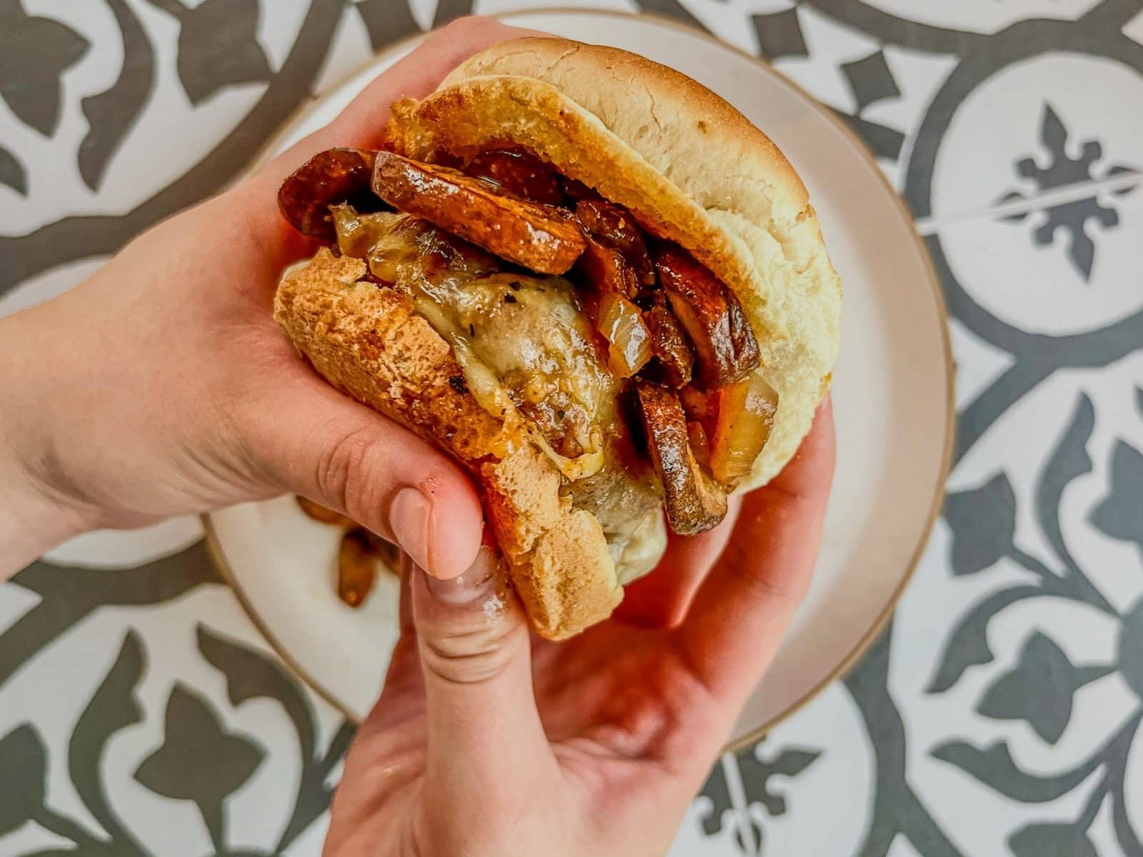 A person holding a burger filled with melted cheese, onions, and grilled vegetables over a patterned plate.