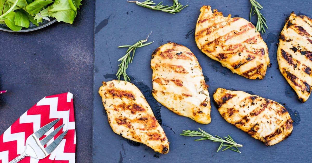 Five grilled chicken breasts with grill marks on a slate, garnished with rosemary, beside salad and a napkin.