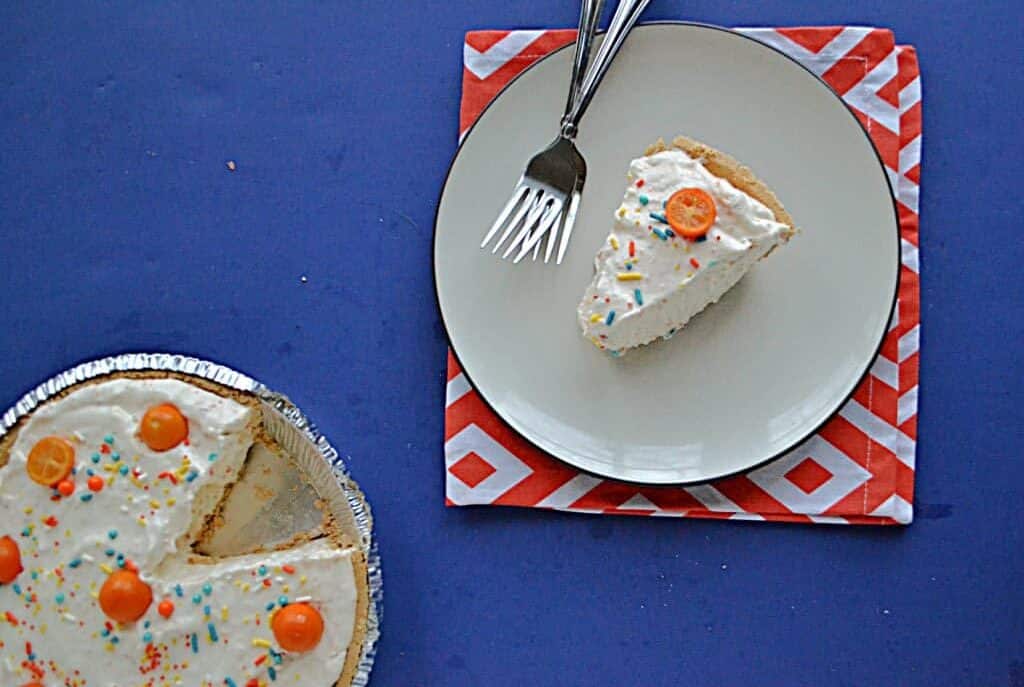 A slice of sprinkled cream pie on a plate with a fork, next to the pie, on a blue and red background.