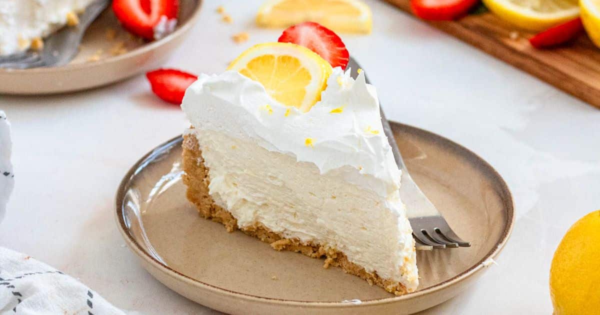 A slice of lemon cheesecake with whipped topping, a lemon slice, and a strawberry on a beige plate with a fork.