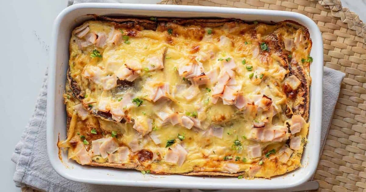 Baked casserole topped with melted cheese and diced ham in a white dish on a woven placemat.