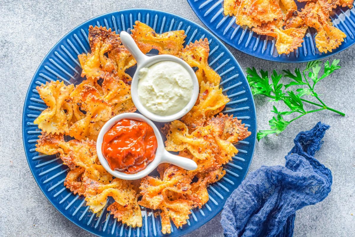 Blue plate with crispy bowtie pasta chips, two dipping sauces (red and white), and a parsley garnish.