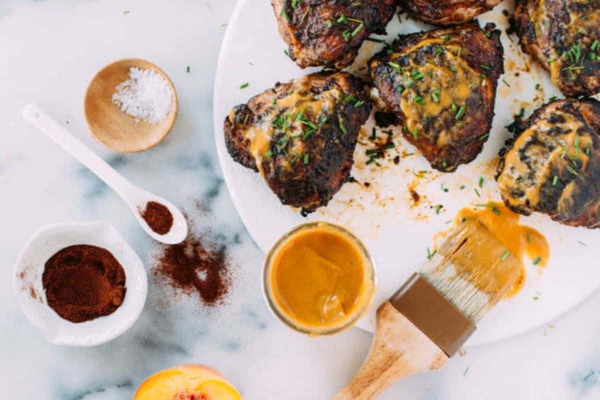 Grilled chicken thighs with sauce, brush, spices, and salt on a marble surface.