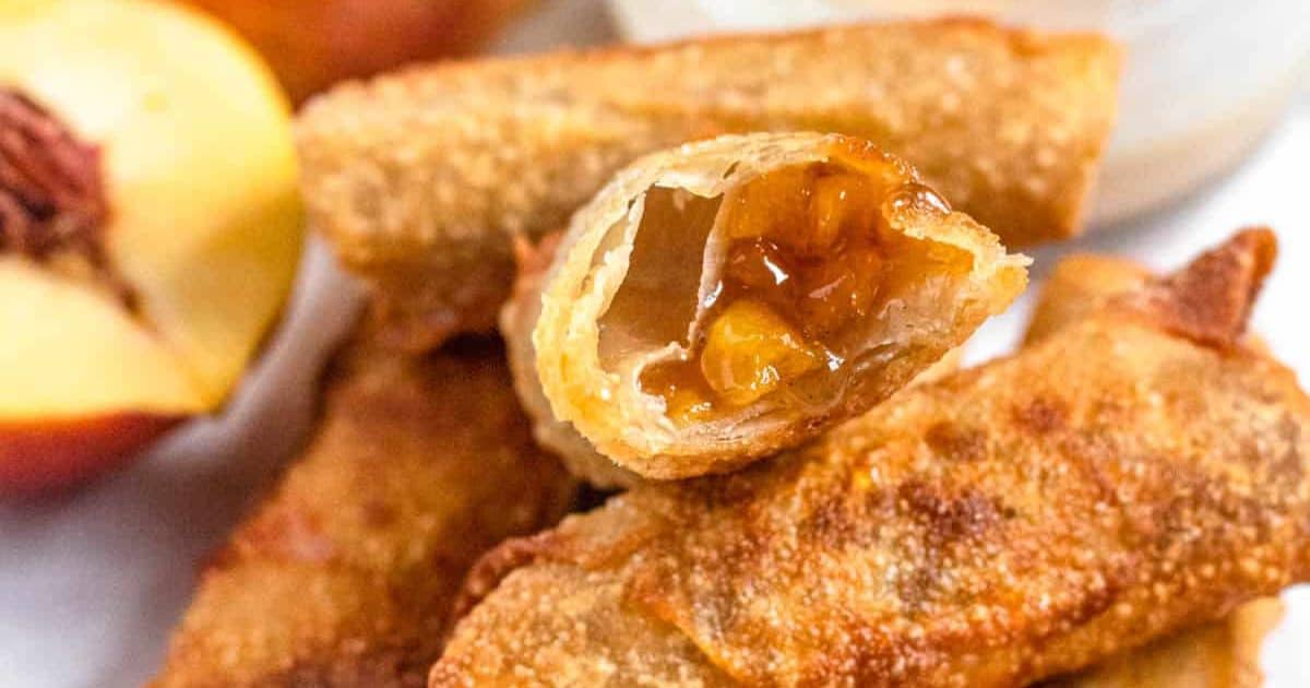 Close-up of crispy peach-filled egg rolls with a halved peach and dipping sauce in the background.