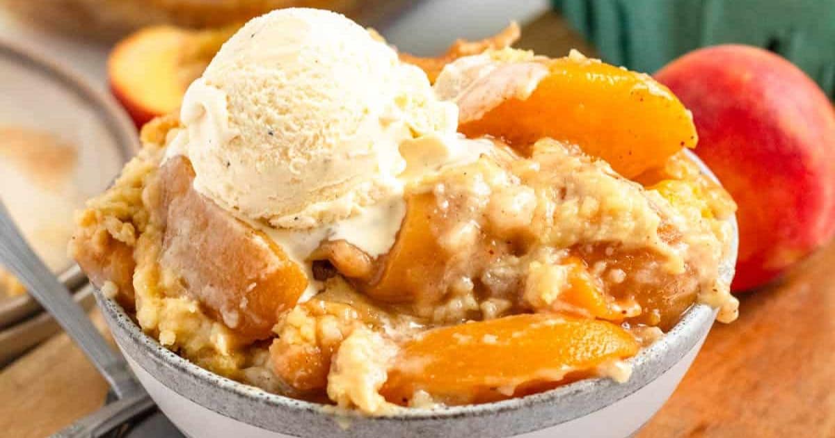 A bowl of peach cobbler topped with a scoop of vanilla ice cream, with fresh peaches in the background.