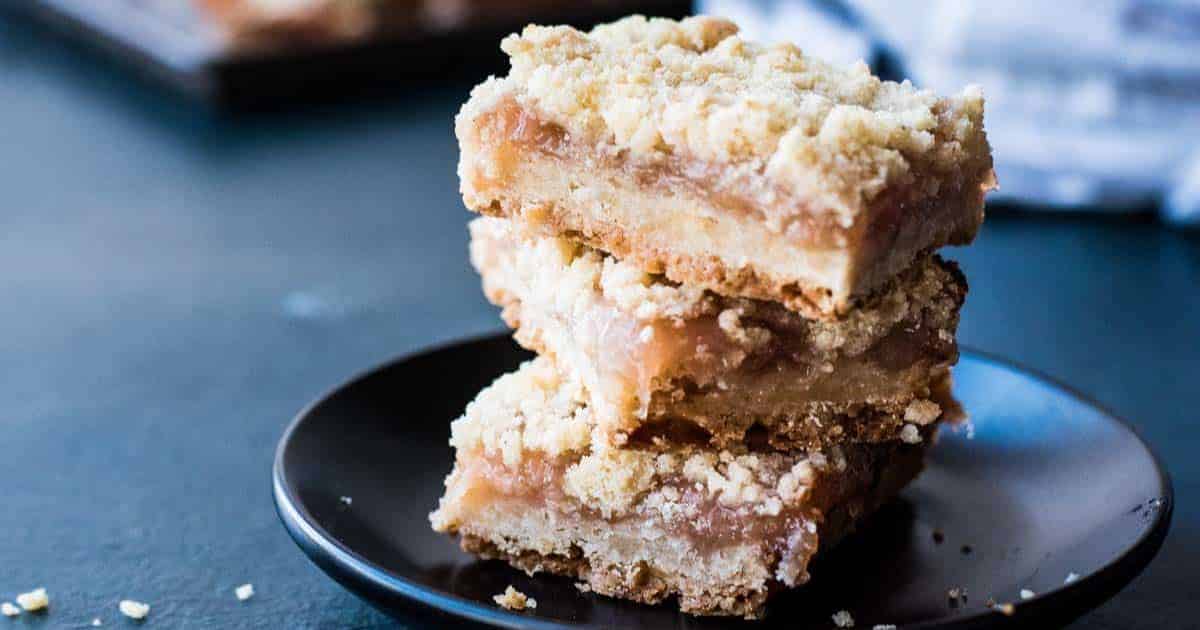 Three crumb-topped dessert bars stacked on a black plate, with a blurred background.