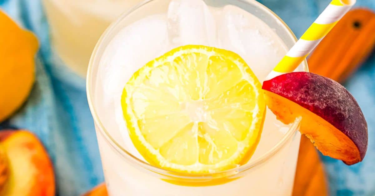 Glass of lemonade with ice, garnished with a lemon slice, peach slice, and a yellow-striped straw.
