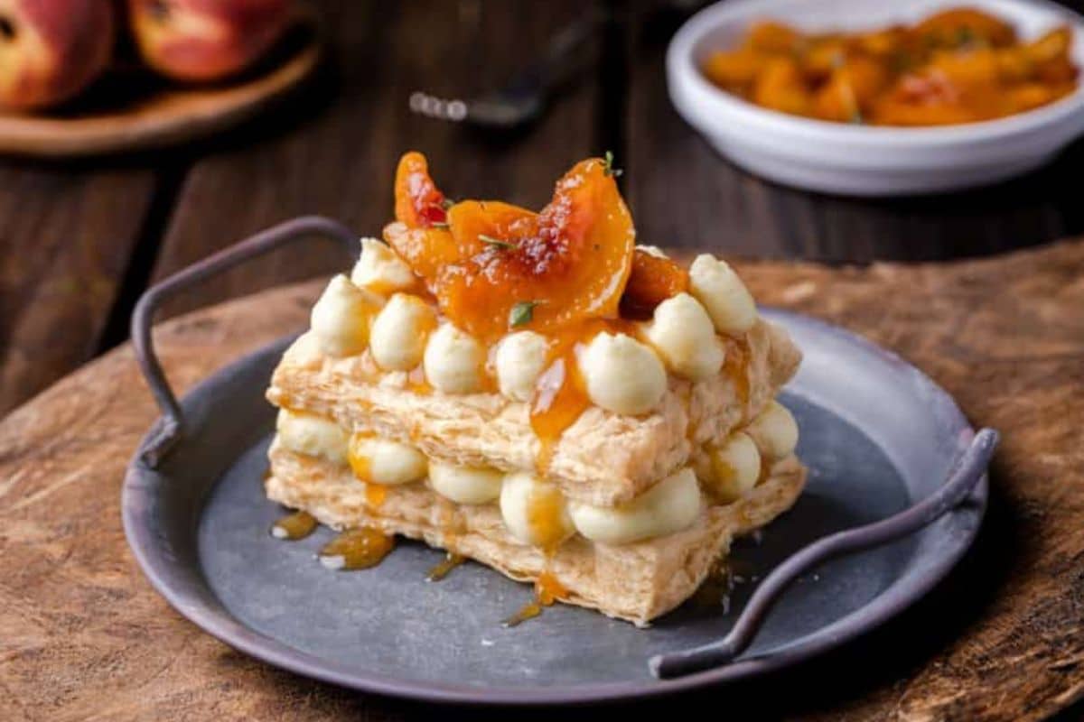 A layered puff pastry dessert with cream filling and caramelized peaches on a metal plate.