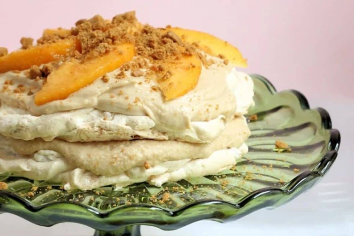 A layered dessert topped with peach slices and crumbled cookies on a green glass cake stand.