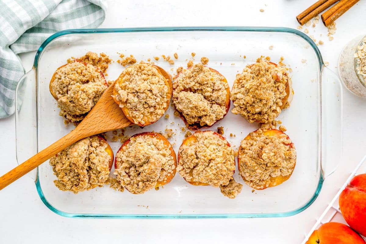 Eight baked apple halves topped with crumbly oat streusel in a glass dish, with a wooden spoon on one.