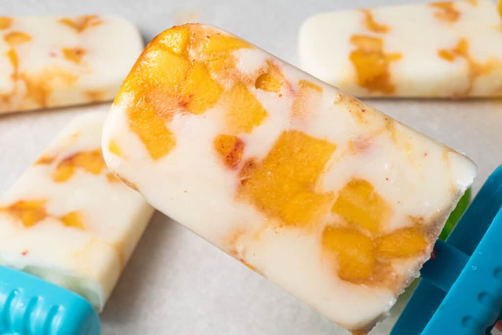 A close-up of a creamy popsicle with visible chunks of yellow and orange fruit, likely mango, on a light surface.