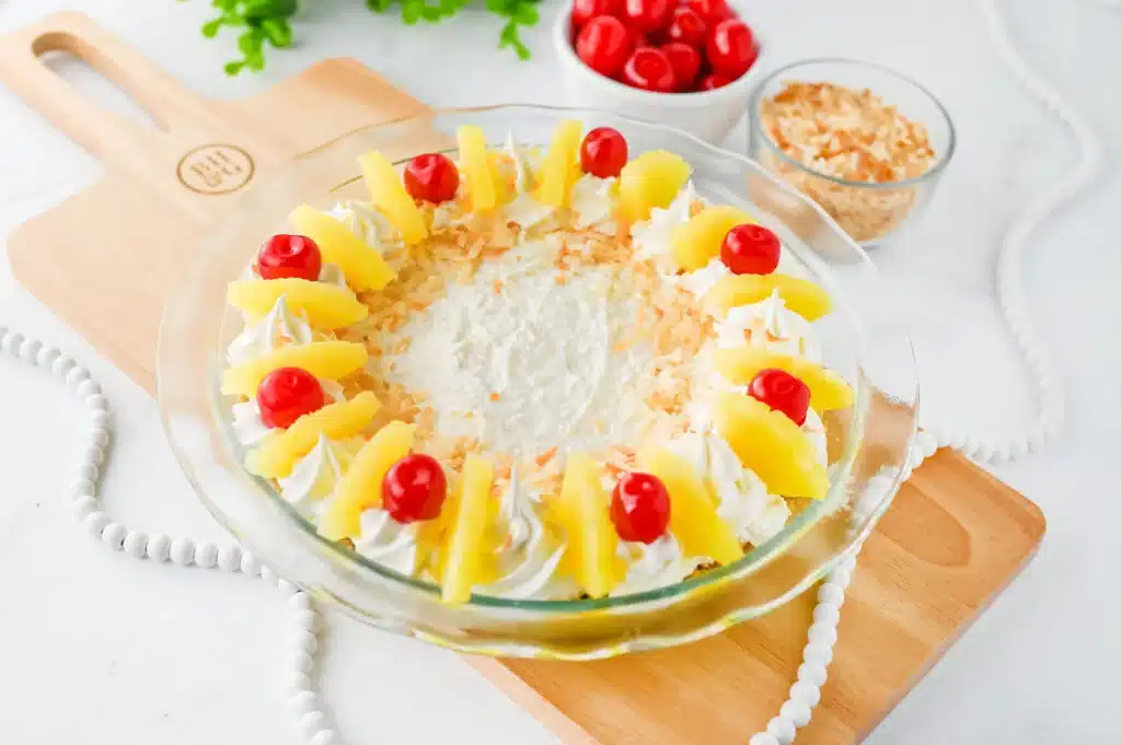 A pie topped with whipped cream, pineapple slices, cherries, and toasted coconut on a wooden board.