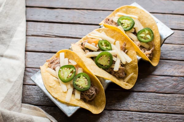 Three corn tacos filled with shredded meat, cheese, and sliced jalapeños on a wooden table.