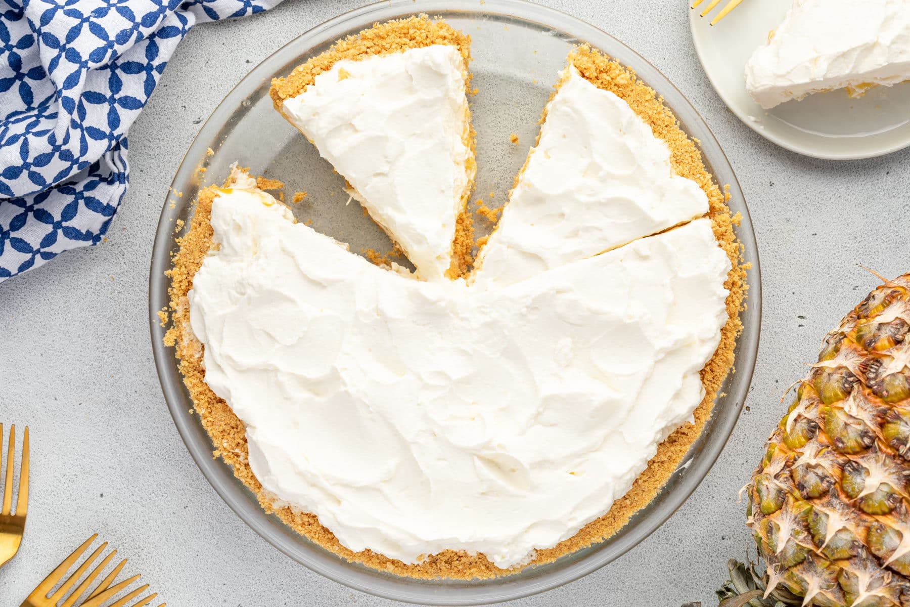 A creamy pie with a crumb crust, two slices removed, on a table with a pineapple and blue napkin nearby.