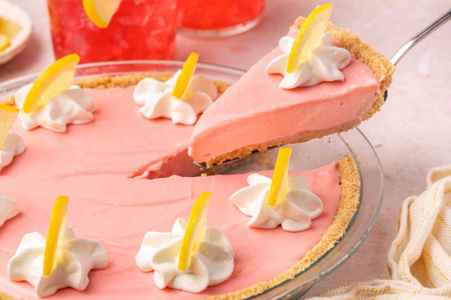A slice of pink cream pie with whipped cream and lemon slices is being lifted from a graham cracker crust pie.