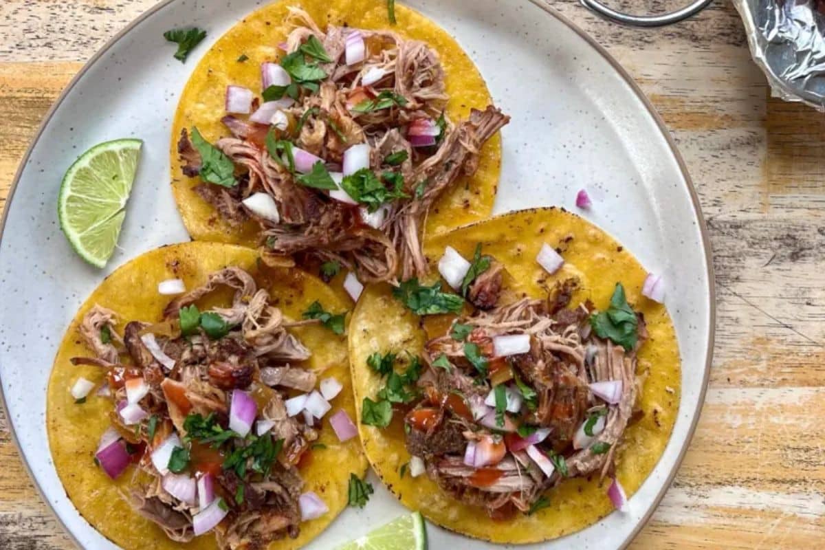 Three tacos with shredded meat, diced onions, and cilantro on yellow tortillas, served with lime wedges on a plate.