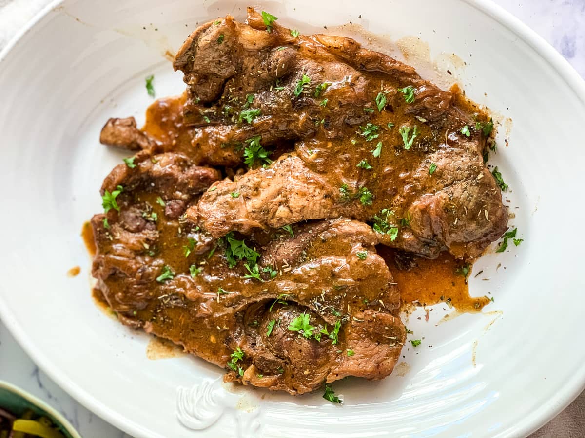 Three pieces of cooked steak with brown gravy, garnished with chopped parsley on a white plate.
