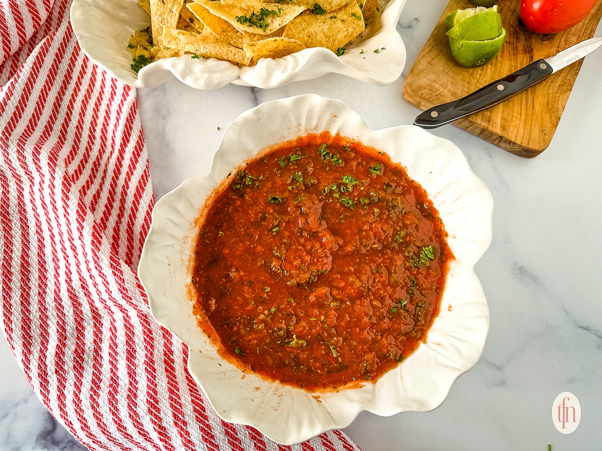 A bowl of chunky red salsa with chopped herbs, next to tortilla chips, lime, and a red-striped towel.