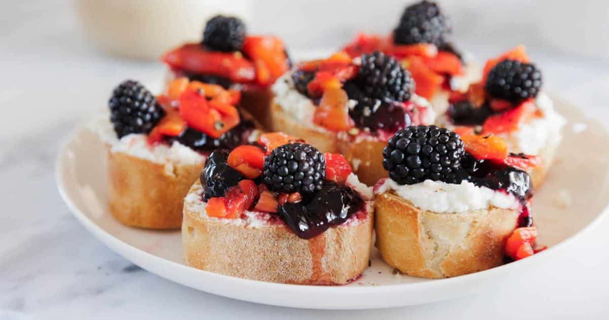 Slices of baguette topped with ricotta, blackberries, and diced red peppers on a white plate.