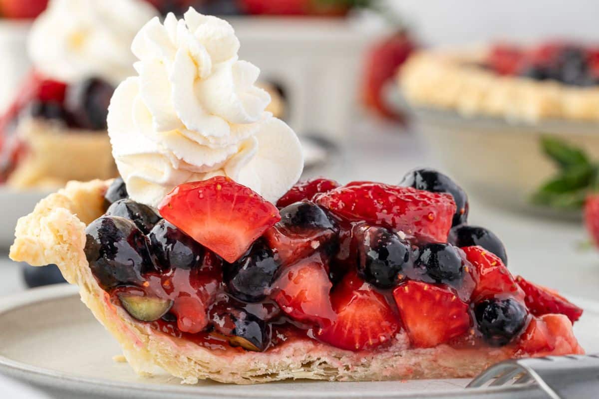 A slice of mixed berry pie topped with whipped cream on a plate, featuring strawberries and blueberries.