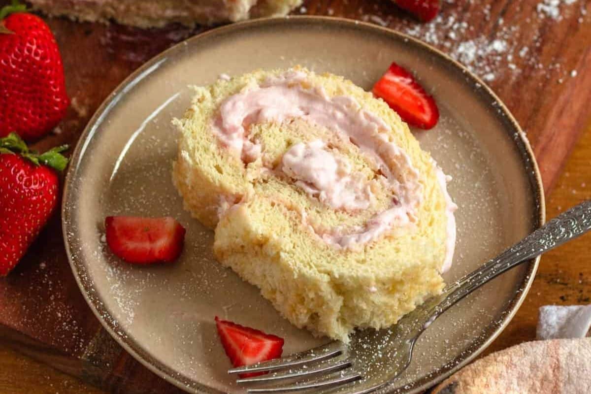 A slice of strawberry cream roll cake on a plate with strawberry pieces and a fork.