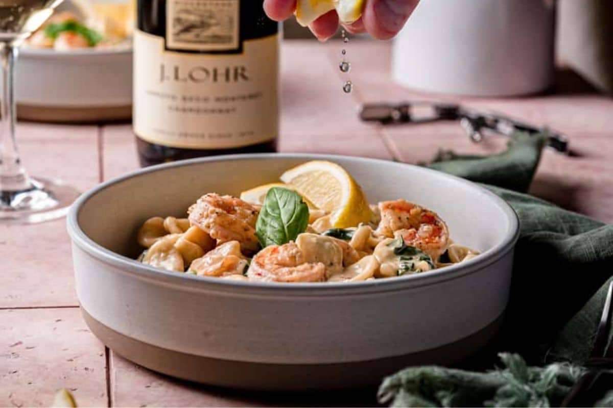 A bowl of shrimp pasta garnished with lemon and basil, with lemon juice being squeezed on top.