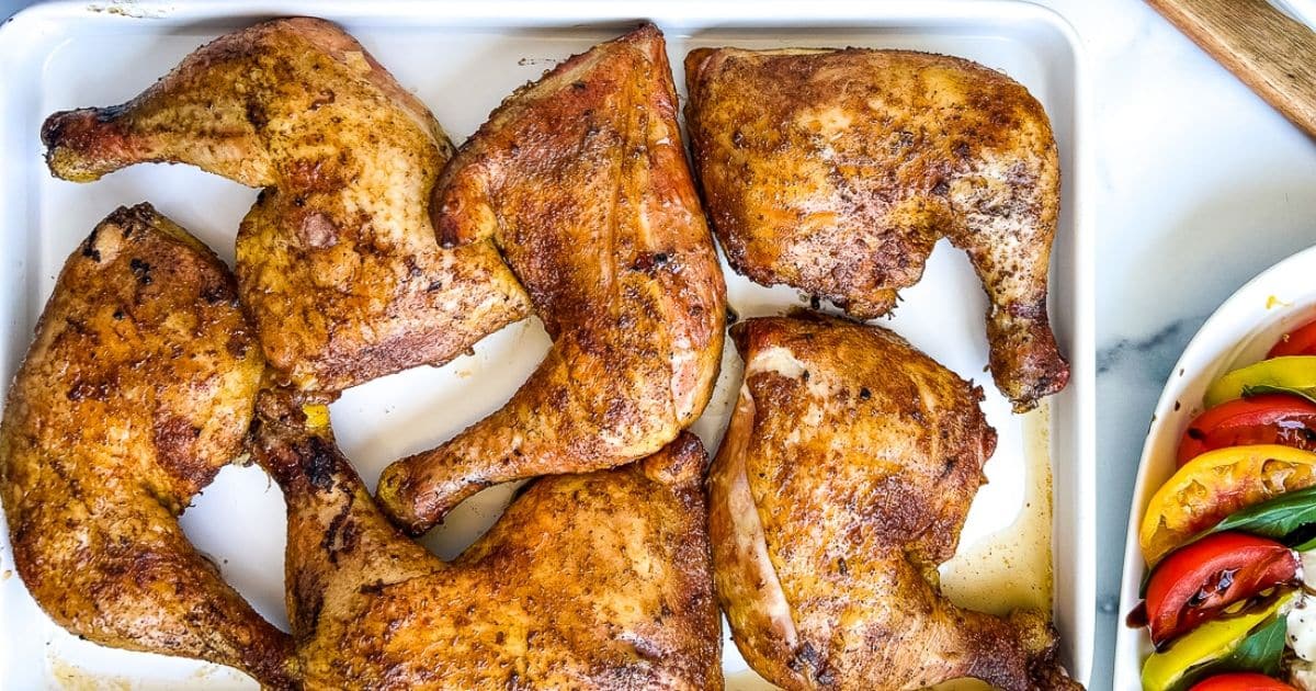 Six seasoned, roasted chicken leg quarters in a white baking dish, with grilled vegetables on the side.
