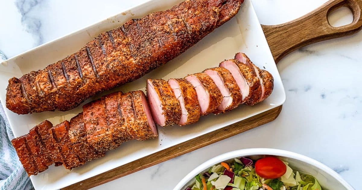 Sliced and whole seasoned pork tenderloin on a white tray, next to a bowl of salad on a marble countertop.