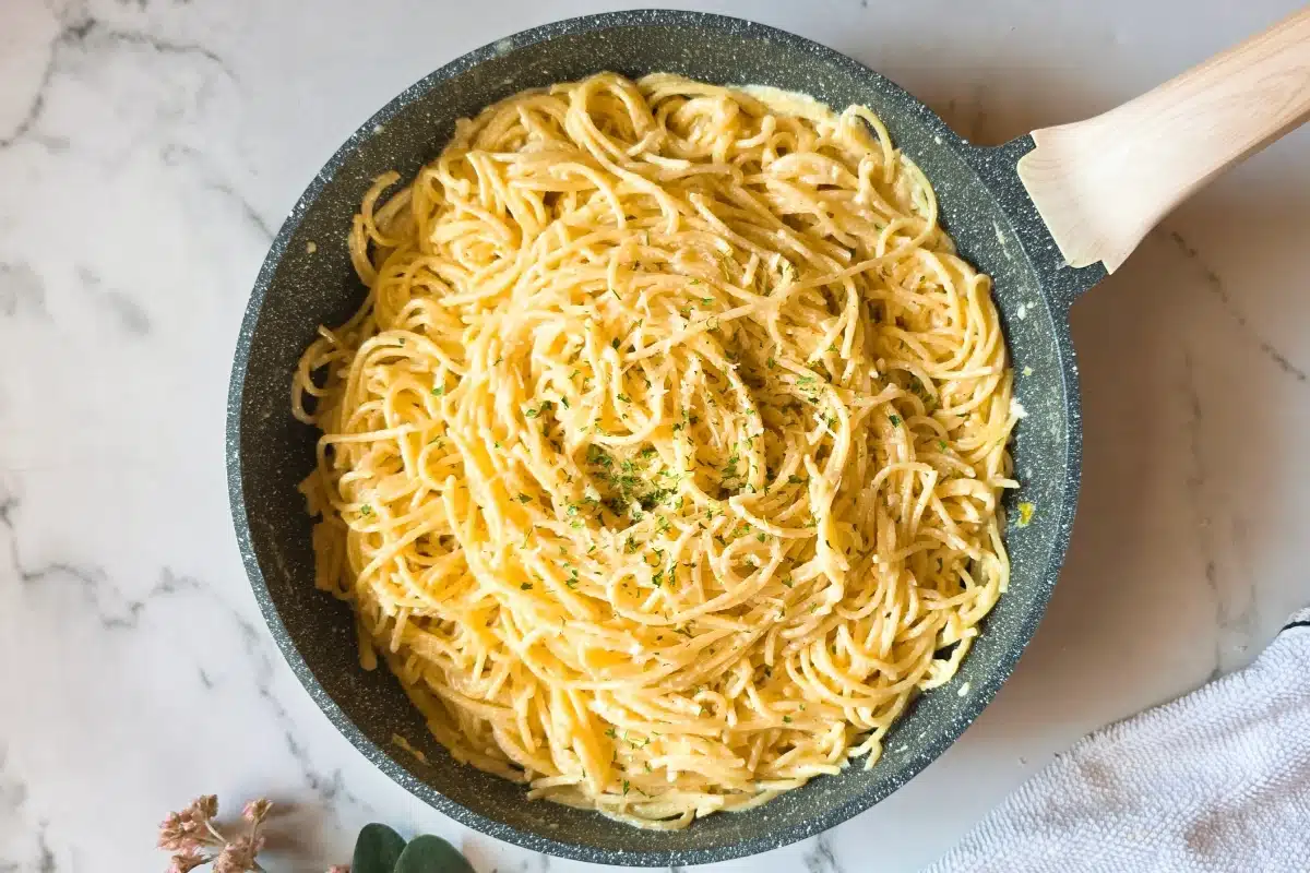 A pan of creamy spaghetti garnished with herbs, placed on a white marble surface.