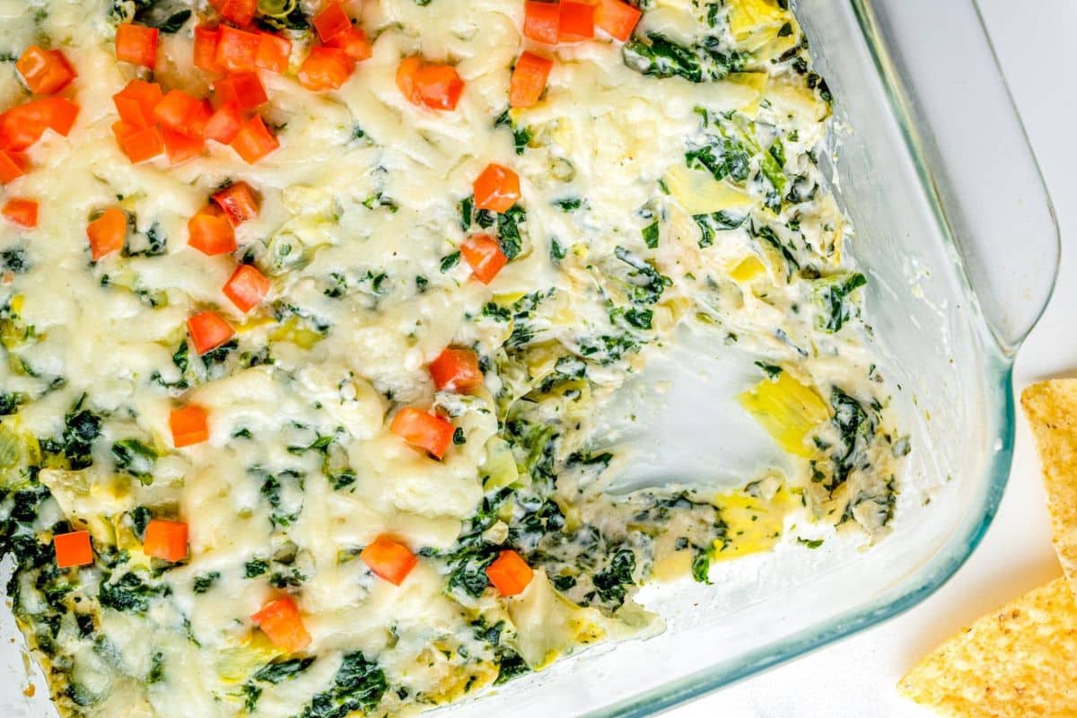 Baked spinach artichoke dip topped with diced tomatoes in a glass dish, some dip removed from the corner.