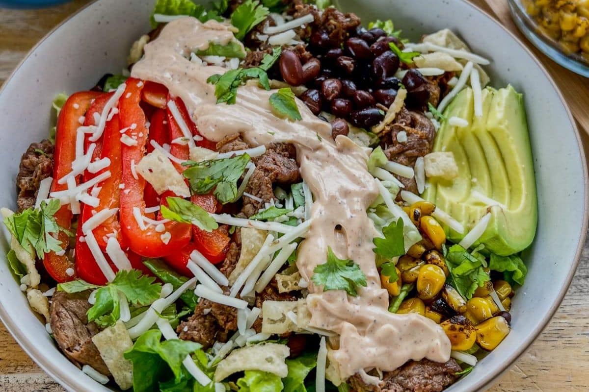 A bowl with lettuce, steak, red peppers, black beans, corn, avocado, cheese, tortilla strips, and dressing.