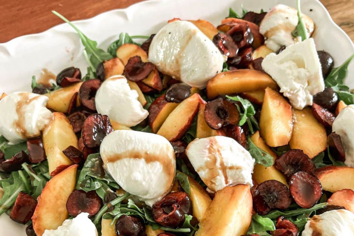 Salad with arugula, peaches, cherries, mozzarella, and balsamic drizzle on a white platter.