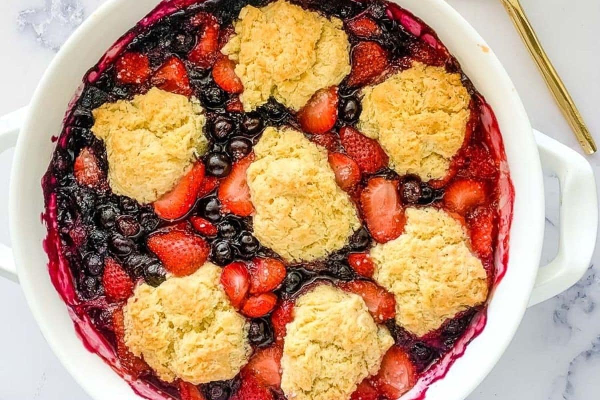Berry cobbler with golden biscuit topping in a white baking dish, showing strawberries and blueberries.