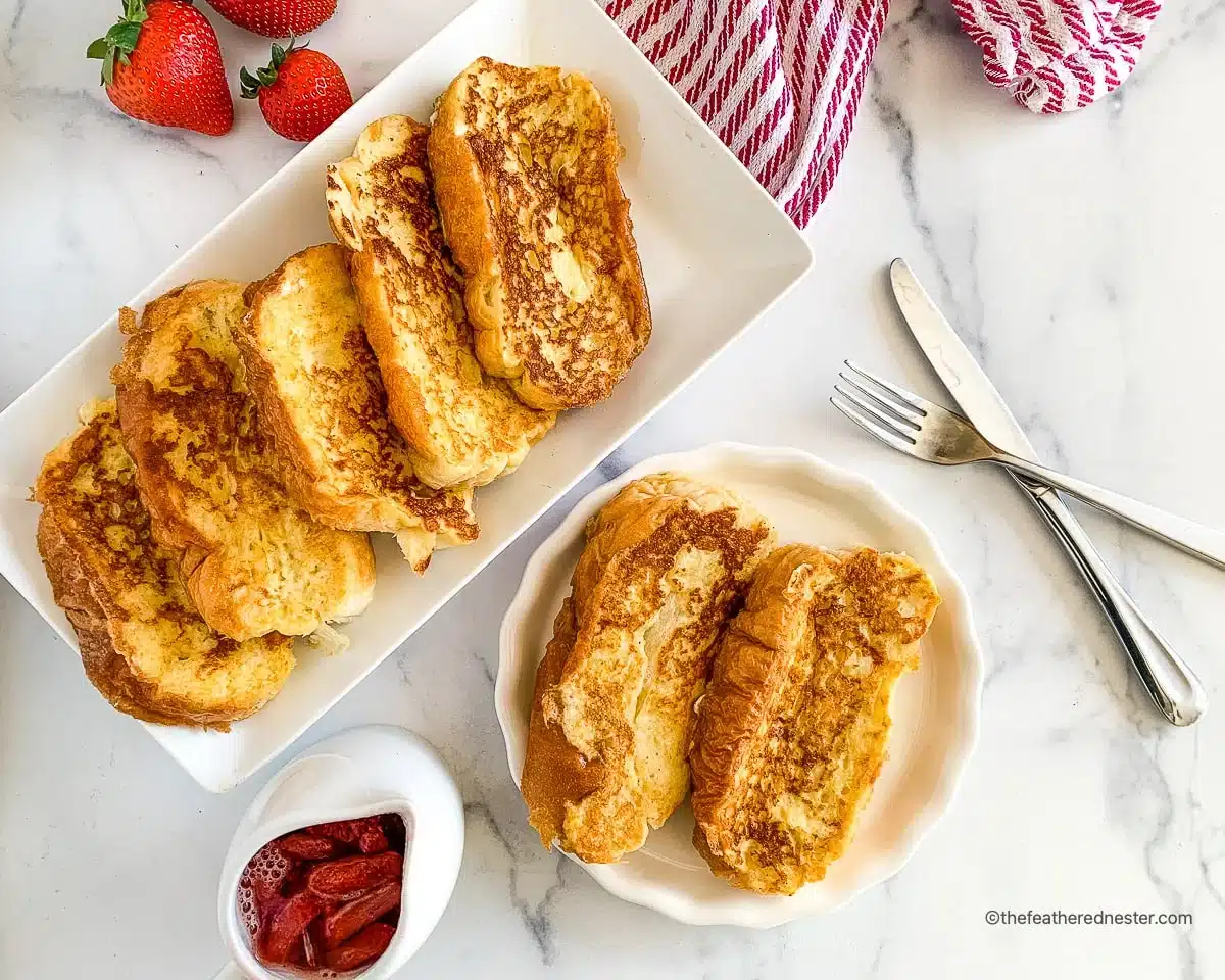 A plate of French toast with strawberries, syrup, fork, and knife on a white marble surface.