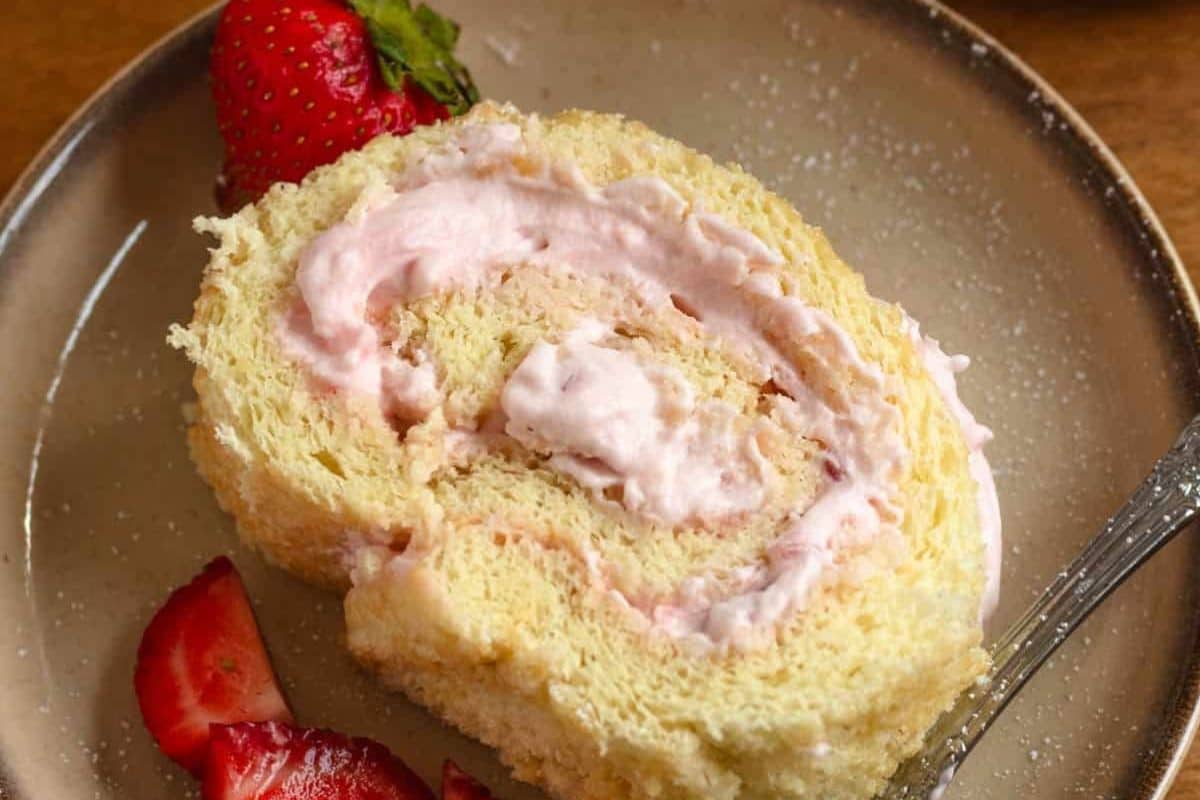 A slice of strawberry cream roll cake on a plate with fresh strawberry slices and a whole strawberry.