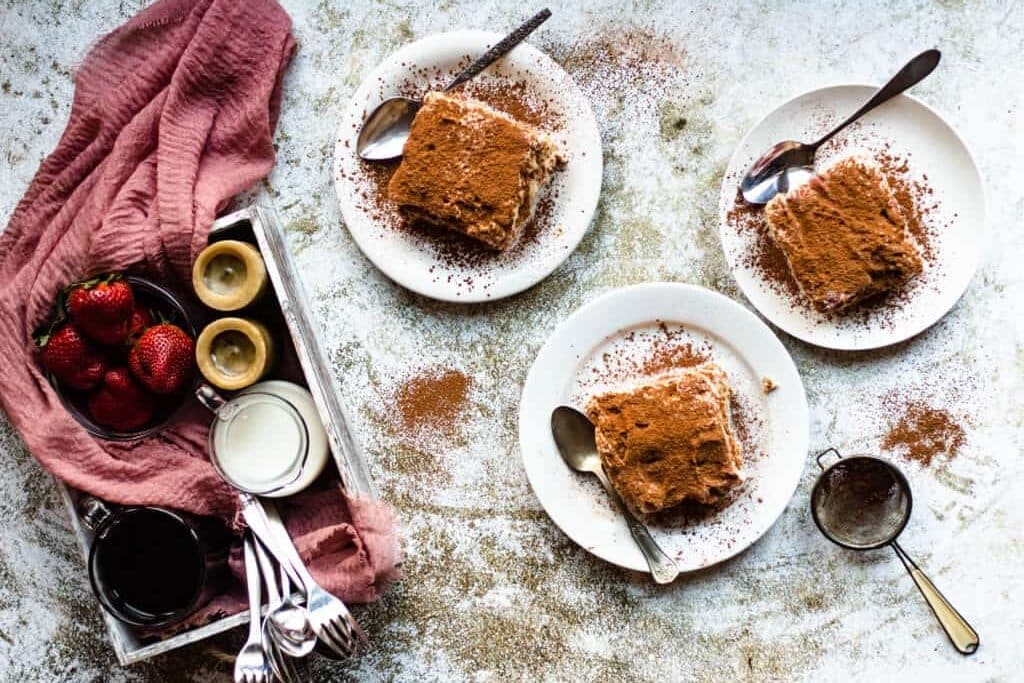 Three plates of tiramisu with spoons, strawberries, and drinks on a rustic surface with a pink napkin.