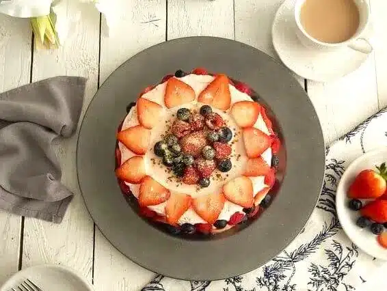 Round cake topped with strawberries and blueberries on a gray plate, with coffee and fruit on a white wooden table.