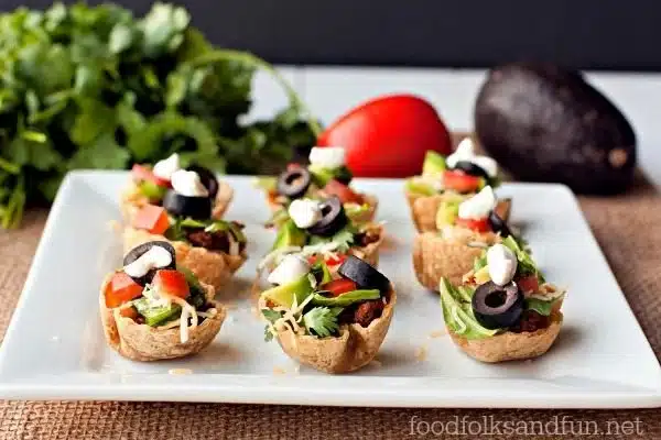 Mini taco cups filled with lettuce, tomato, olives, cheese, and sour cream on a white plate.