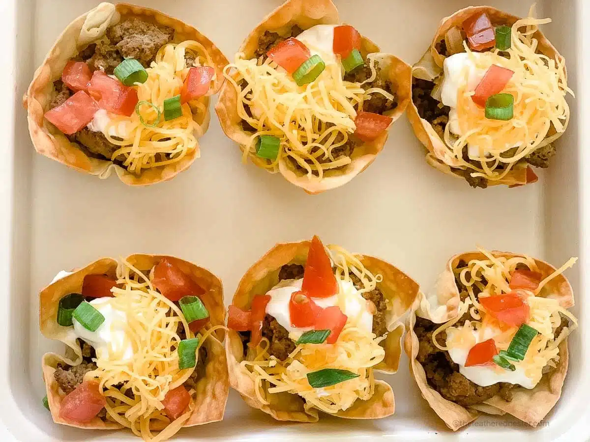 Six taco cups with ground beef, shredded cheese, sour cream, tomatoes, and green onions in a baking dish.