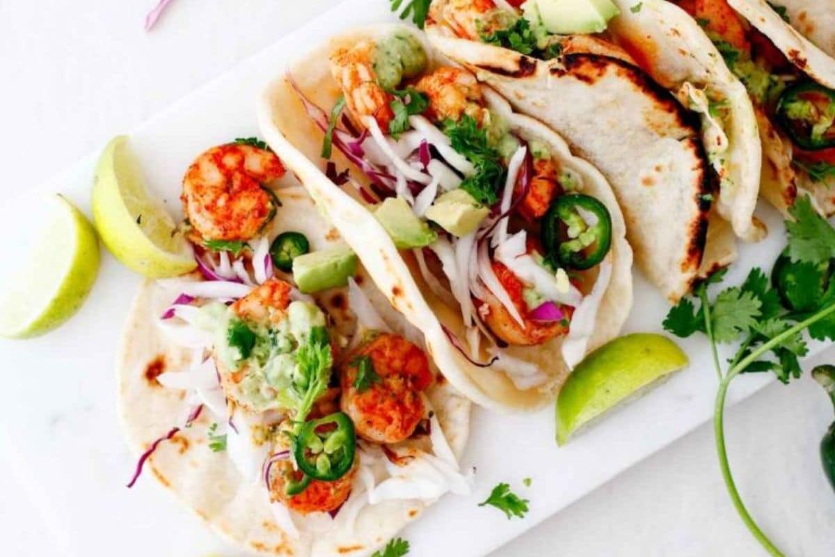 Three shrimp tacos with cabbage, avocado, jalapeños, and cilantro on tortillas, served with lime wedges.