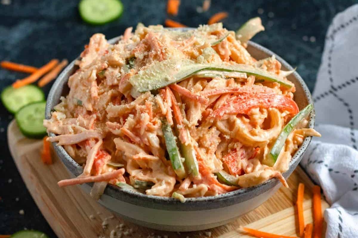 A bowl of creamy coleslaw with shredded carrots, cucumbers, and green peppers on a wooden board.