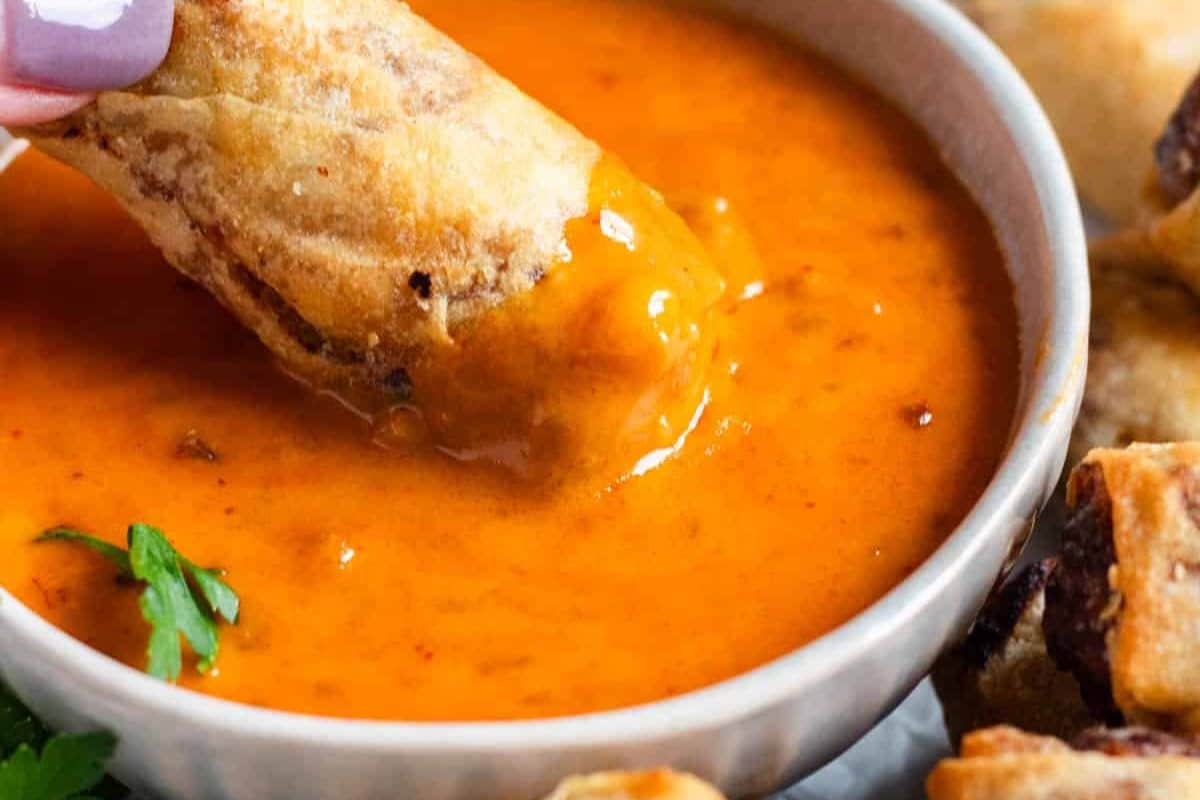 A hand dips a crispy spring roll into a bowl of bright orange dipping sauce.