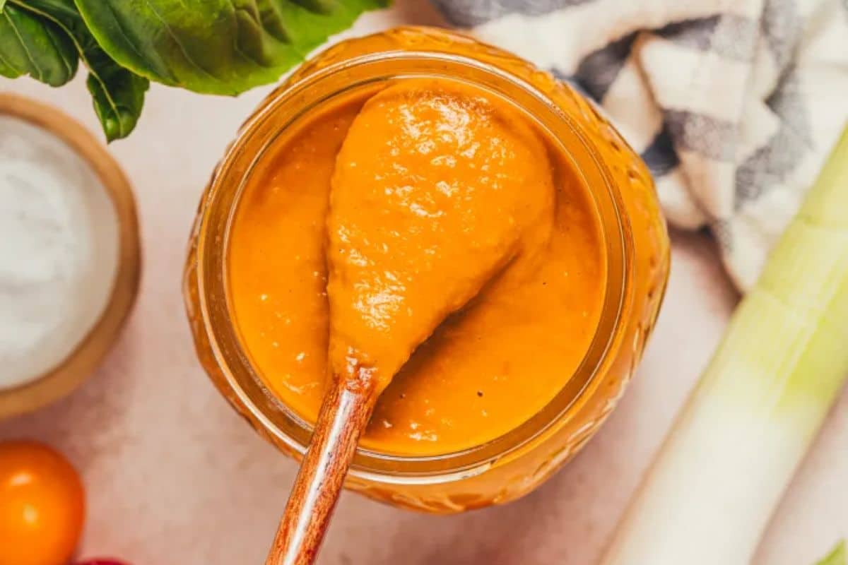 A wooden spoon in a jar of orange tomato sauce, surrounded by fresh vegetables and a towel.